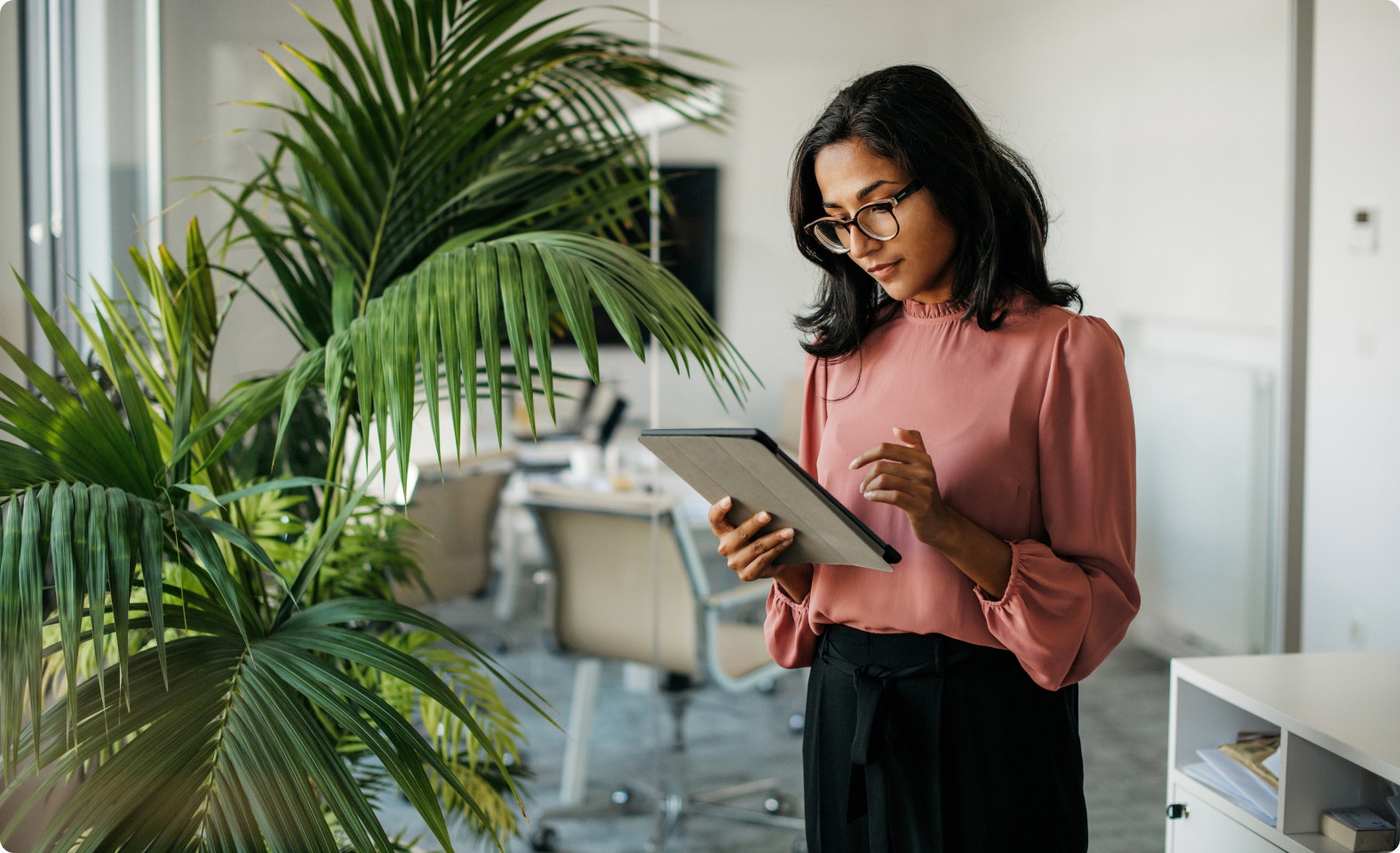 Woman standing near an office window analyzing a report on a tablet.